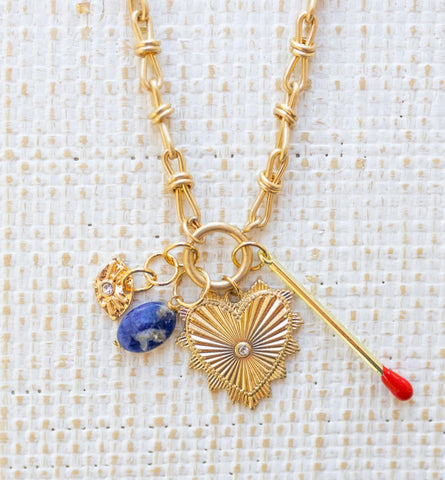 Picture of gold toggle chain with a large gold heart pendant with a diamond at the center. Necklace has three additional charms, a gold and diamond evil eye, a blue marbled bead and a gold matchstick.