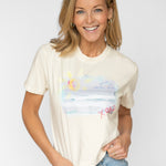 Happy blonde woman wearing a cream colored t shirt tucked into her blue jeans. T shirt has a beach scene printed on the front of it with the letters XOXO in pink.
