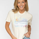 Happy blonde woman wearing a cream colored t shirt tucked into her blue jeans. T shirt has a beach scene printed on the front of it with the letters XOXO in pink.