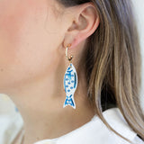 Picture of a woman wearing a small gold hoop with a dangling white fish bead with blue details.