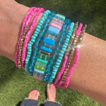 Picture of a woman's wrist displaying a beautiful set of bracelets. Six pink and gold small bead bracelets, 2 solider blue bracelets, 2 clear blue bracelets and one thicker beaded bracelet made of rectangular beads in blues, greens and pinks.