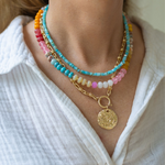 Woman wearing three necklaces: a gold chunky chained necklace sitting on a table with a large gold medallion attached to it, a multi-colored stone beaded short necklace, and a small blue beaded choker