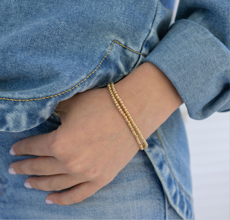 Model's wrist displaying two tiny gold bead bracelets.