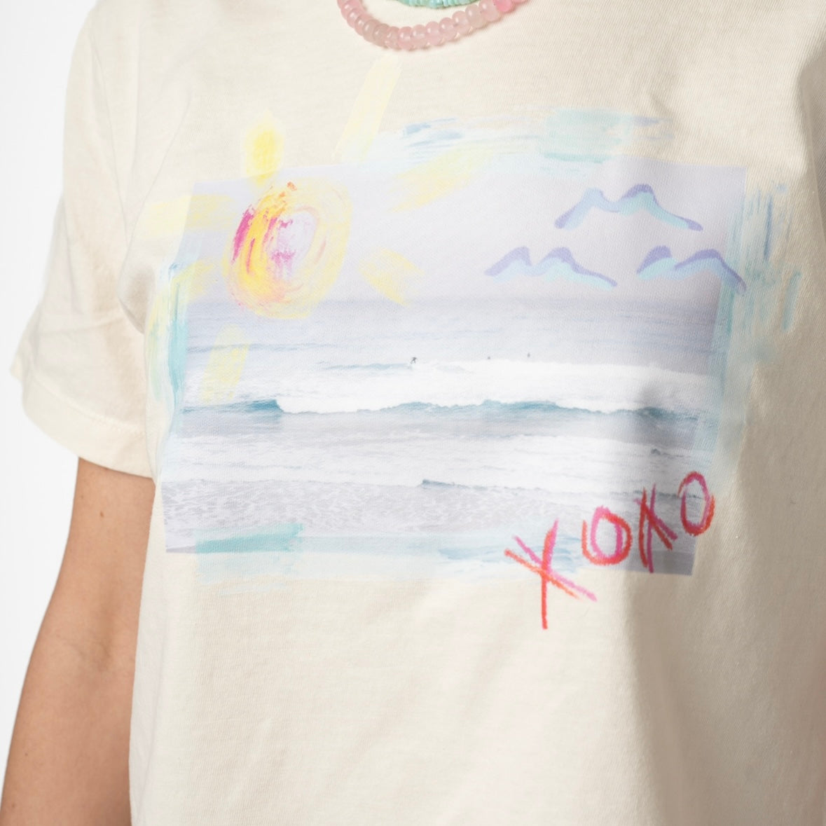 Picture of a woman wearing a cream colored Tshirt with a beach scene printed on it. It also has the letters XOXO written on it. She is wearing a semi-precious stone beaded necklace in light pink, yellow and teal, layered with a tiny teal beaded necklace.