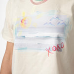 Picture of a woman wearing a cream colored Tshirt with a beach scene printed on it. It also has the letters XOXO written on it. She is wearing a semi-precious stone beaded necklace in light pink, yellow and teal, layered with a tiny teal beaded necklace.