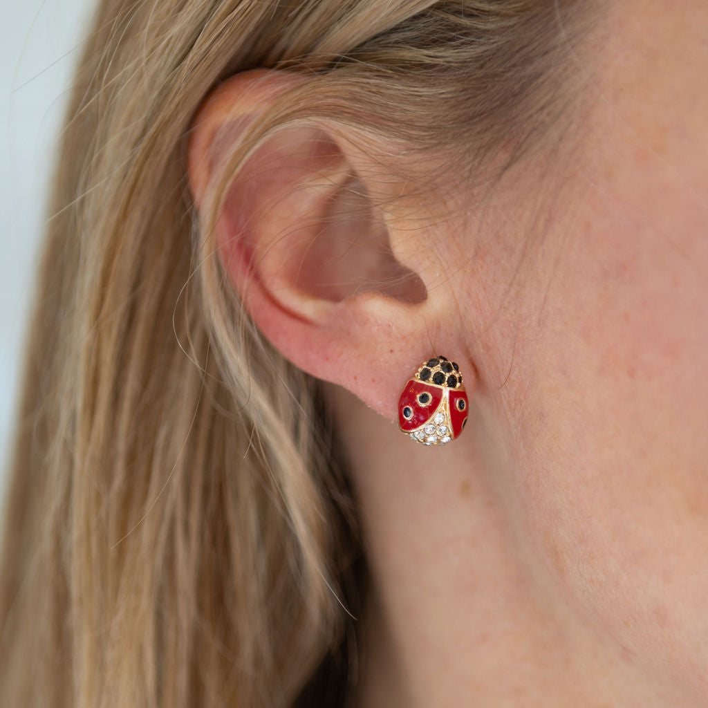 A person wearing ladybug stud earrings with red and black enamel design and a small quartz stone.