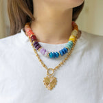 Model wearing large multicolored beaded necklace stacked with front clasping gold chain necklace with a heart pendant