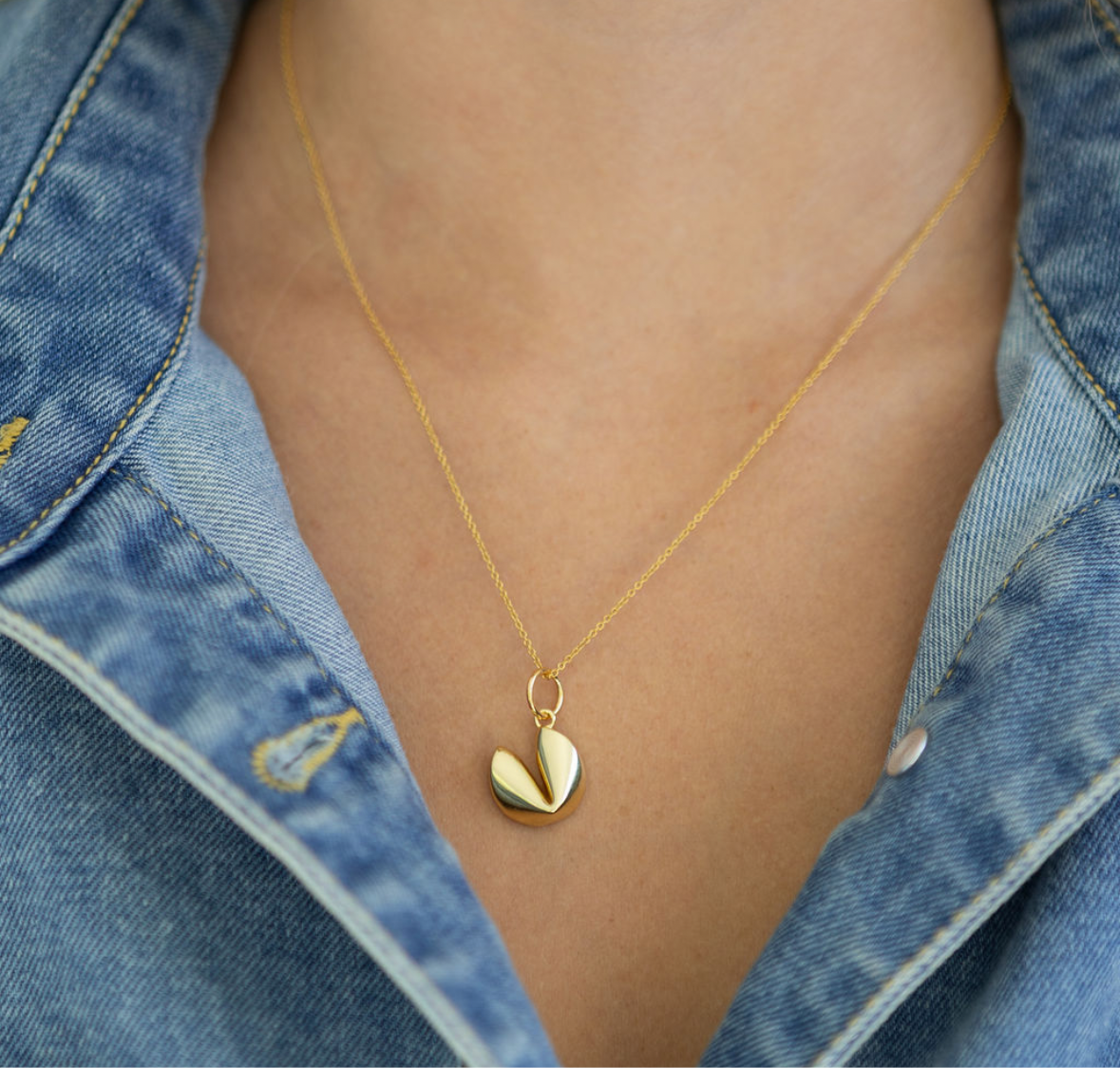 Gold necklace with a gold fortune cookie pendant worn by a person wearing a denim jacket.