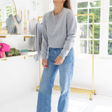 Woman standing in a house posing wearing a gray sweatshirt with blue stitching that says  "Joie De Vivre"