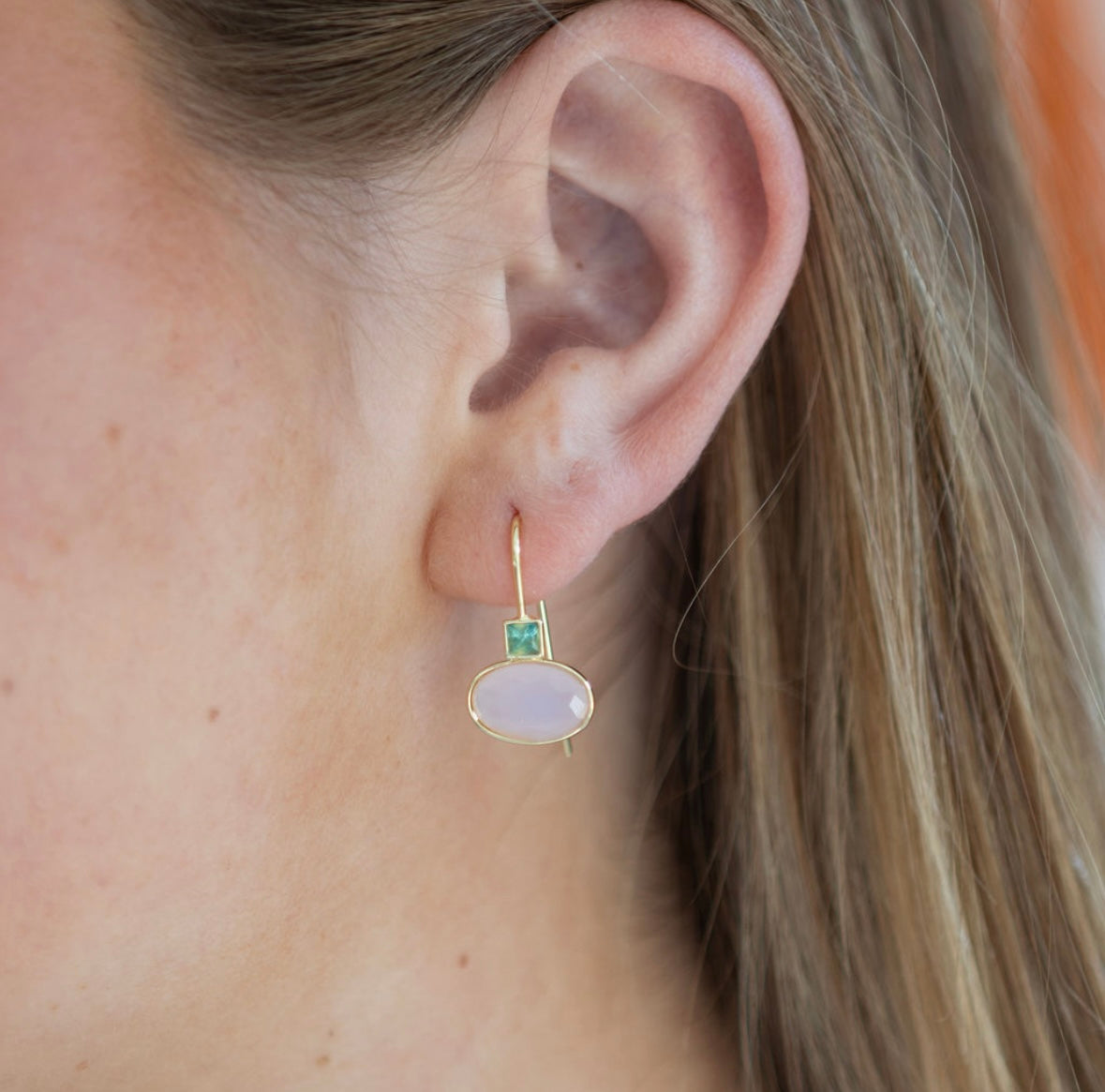 Close-up of an ear wearing a gold earring with a large light pink oval gemstone and a tiny square green gemstone.