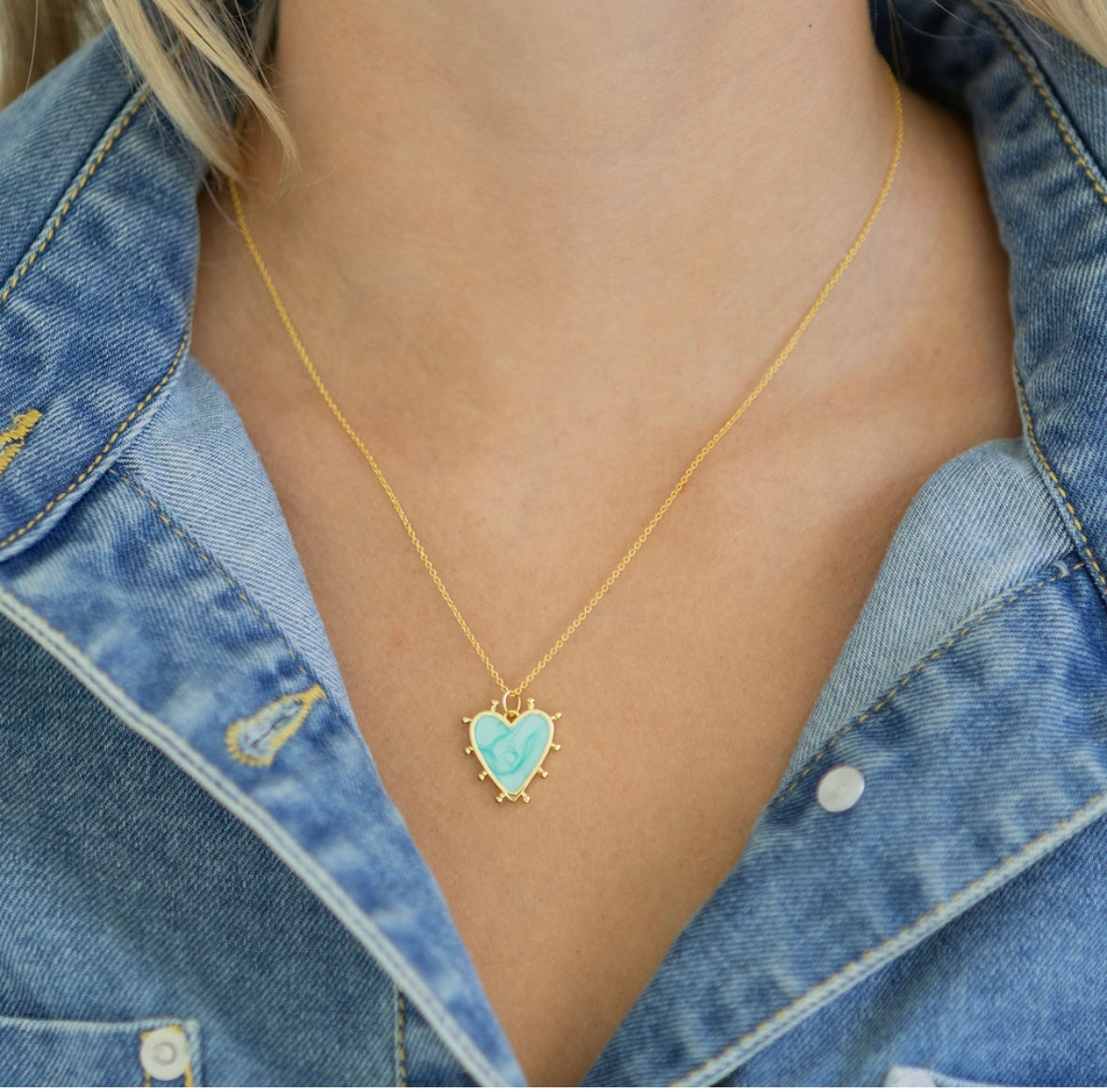 Gold necklace with a turquoise heart pendant worn by a person in a denim jacket.