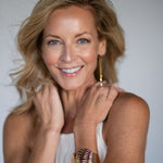 Woman with blonde hair and a white top, wearing a gold clam shell cuff bracelet and gold dangling earrings, against a plain background