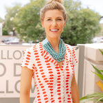Woman outside smiling wearing a white dress with a red lips print and a big blue stone layered necklace around her neck