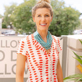 Woman outside smiling wearing a white dress with a red lips print and a big blue stone layered necklace around her neck