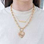 Model wearing a fine gold chain necklace with bar pendant and large gold chain charm necklace with gold heart pendant with diamond at its center.