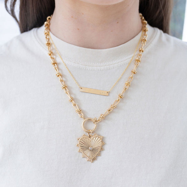 Model wearing a fine gold chain necklace with bar pendant and large gold chain charm necklace with gold heart pendant with diamond at its center.