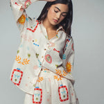 Picture of a woman wearing a white silk button up top with matching pants. Clothing set has bright shapes and plant and sun themed pictures. 