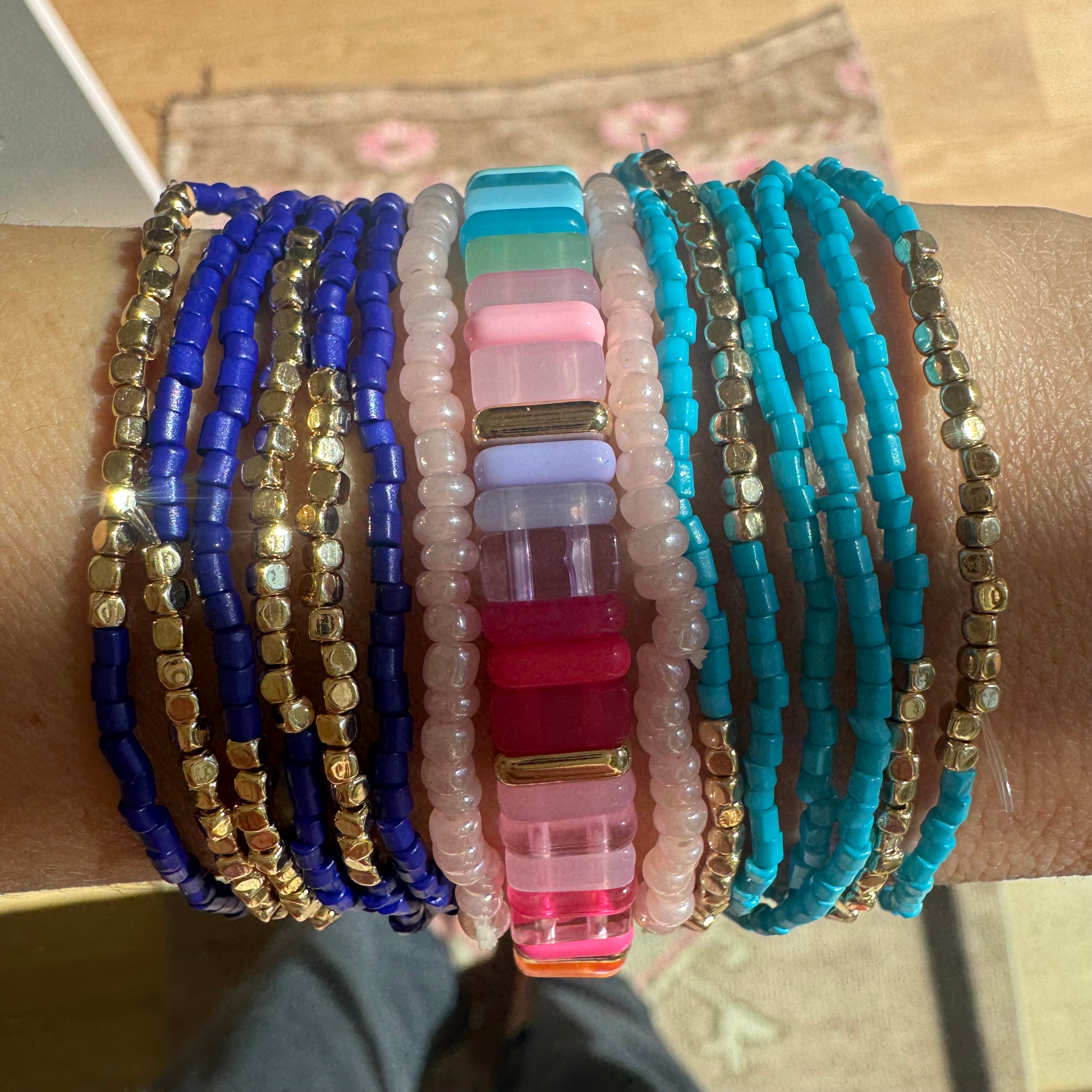 Picture of a set of bracelets. 6 dark blue and gold bracelets, 2 pearl rose pink bracelets, 6 light blue and gold bead bracelets and one larger bracelet made out of rectangle beads in shades of pinks and blues.