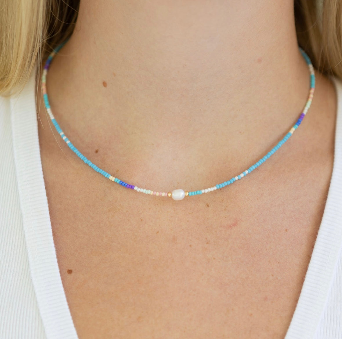 Necklace with small blue and white beads and a pearl on a person wearing a white top.