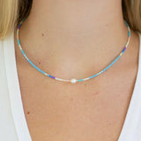Necklace with small blue and white beads and a pearl on a person wearing a white top.