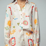 Picture of a woman wearing a white silk button up top with matching pants. Clothing set has bright shapes and plant and sun themed pictures. 
