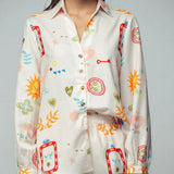Picture of a woman wearing a white silk button up top with matching pants. Clothing set has bright shapes and plant and sun themed pictures. 