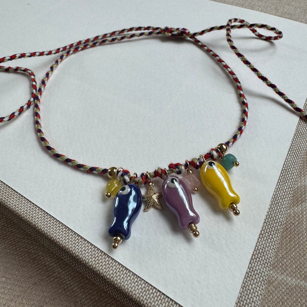 Picture of a thin woven  adjustable string bracelet in red, white and black. Dangling from it are three fish beads in blue, purple and yellow with small yellow, pink and green accent beads and a small gold starfish.