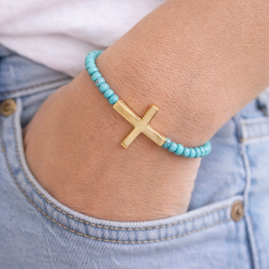 Turquoise beaded bracelet with a gold cross on a wrist, wearing light blue jeans.