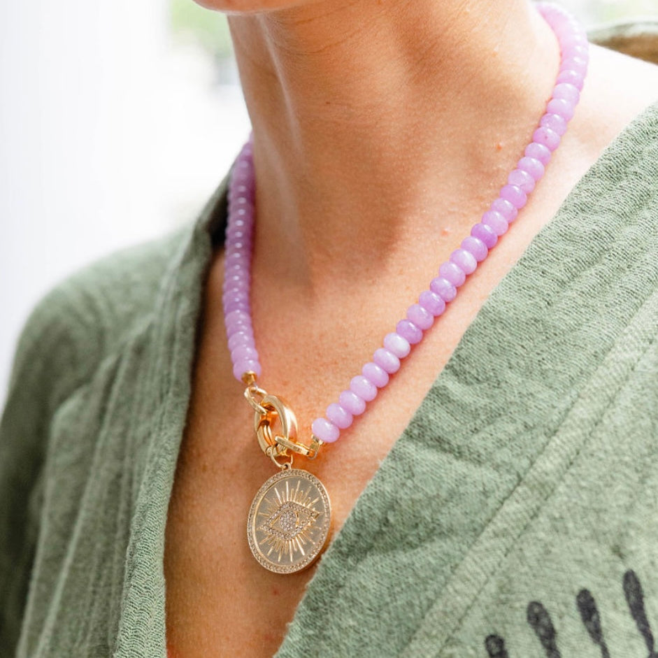 A girl wearing a light pink stone beaded necklace with a gold chain link in the middle with a gold engraved eye medallion attached to it