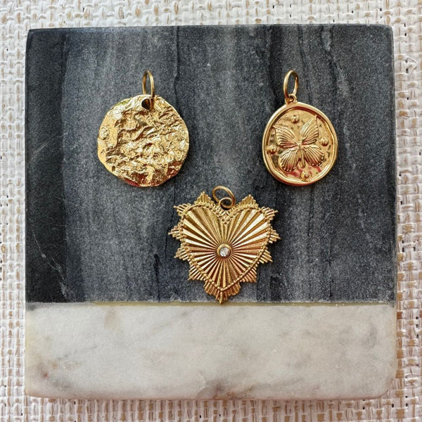 Picture of three gold pendants. The first is a hammered gold disc, second a gold disc with a butterfly design and the third a textured gold heart with a clear stone at its center.