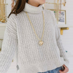 Woman in a white turtleneck wearing a long gold chain with a gold Puffy heart pendant.