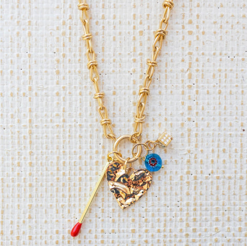 Picture of gold toggle chain with a textured gold heart pendant. Three additional charms attached include gold matchstick, blue bead and gold dice.