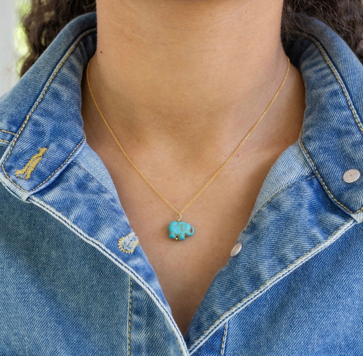 Person wearing a gold necklace with a turquoise elephant pendant and a denim jacket.