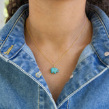 Person wearing a gold necklace with a turquoise elephant pendant and a denim jacket.