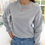 Woman standing in a house posing wearing a gray sweatshirt with blue stitching that says  "Joie De Vivre"