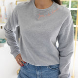 Woman standing in a house posing wearing a gray sweatshirt with blue stitching that says  "Joie De Vivre"