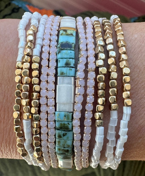 Picture of a woman's wrist displaying 11 bracelets. 6 bracelets are dainty white and gold beads, 4 bracelets are white quartz beads and the main bracelet is light mottled turquoise and mother of pearl beads.