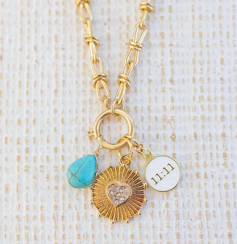 Picture of gold toggle chain with various charms attached.  One is a turquoise tear drop charm, gold textured disc with diamond heart in the center and white 11:11 disc.