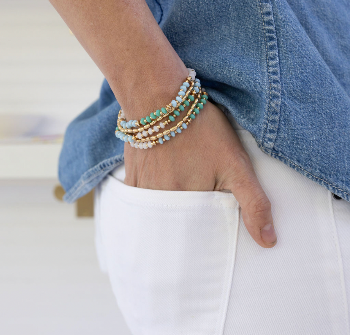 Hand wearing multiple bracelets with white beads, teal beads and gold beads with a denim jacket and white pants