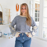 Woman wearing a blue and white patterned sweater with floral cuffs in a room with jewelry and decor.