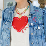 Woman wearing a denim jacket with embroidered hearts and a cream colored undershirt with a large red heart and red script that reads "love". Model is also wearing a colorful bead necklace and gold chain necklace with a heart pendant.