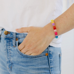 Picture of a woman with her hand in her pocket. She is wearing a beaded bracelet in yellows, purples, reds and blues.