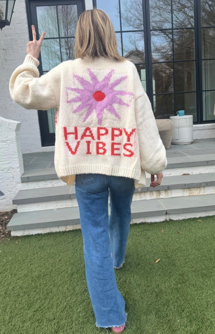 Picture of a woman walking away, holding up her hand in a peace sign, wearing an oversized cream colored sweater with a large purple sunburst with a red center and the words Happy Vibes in red at the bottom.