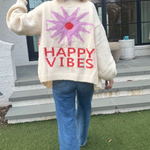 Picture of a woman walking away, holding up her hand in a peace sign, wearing an oversized cream colored sweater with a large purple sunburst with a red center and the words Happy Vibes in red at the bottom.