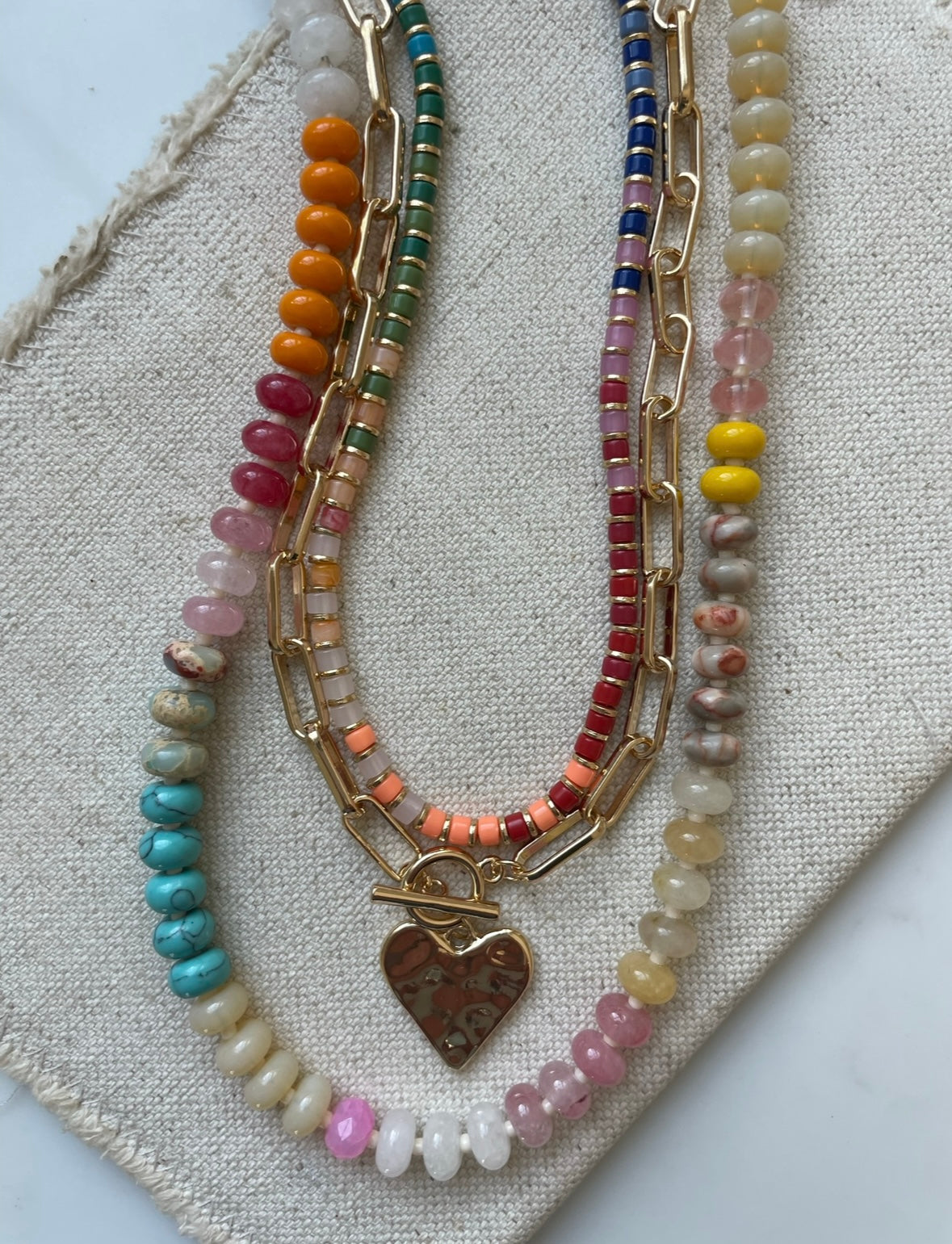 Set of three necklaces made up of small colorful bead necklace, a gold chain link with hammered gold heart pendant and a larger colorful beaded necklace.