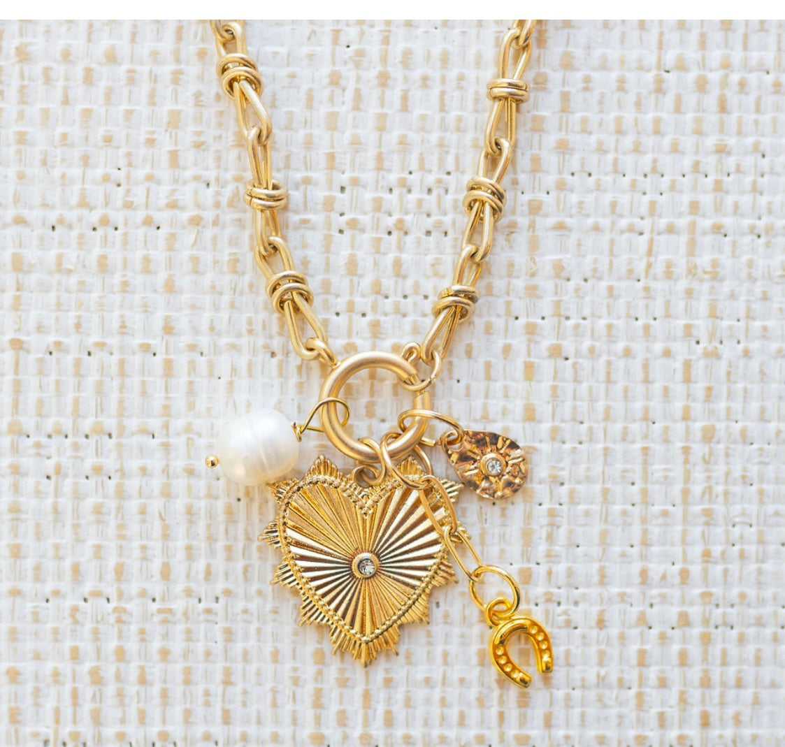 Picture of gold toggle chain with a large gold heart pendant with a diamond in the center. Three additional charms are attached. One is a fresh water pearl, one a gold horseshoe and one a gold teardrop with a small diamond at the center.