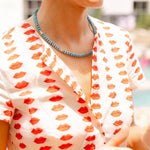 Woman outside wearing a white dress with a red lips print and a blue/gold beaded necklace around her neck