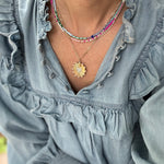 Picture of a woman wearing a blue blouse and four necklaces. 3 necklaces are dainty bead necklaces in pink and white, blue and light blue and white and gold. One necklace is a gold link chain necklace with a large textured gold disc pendant.