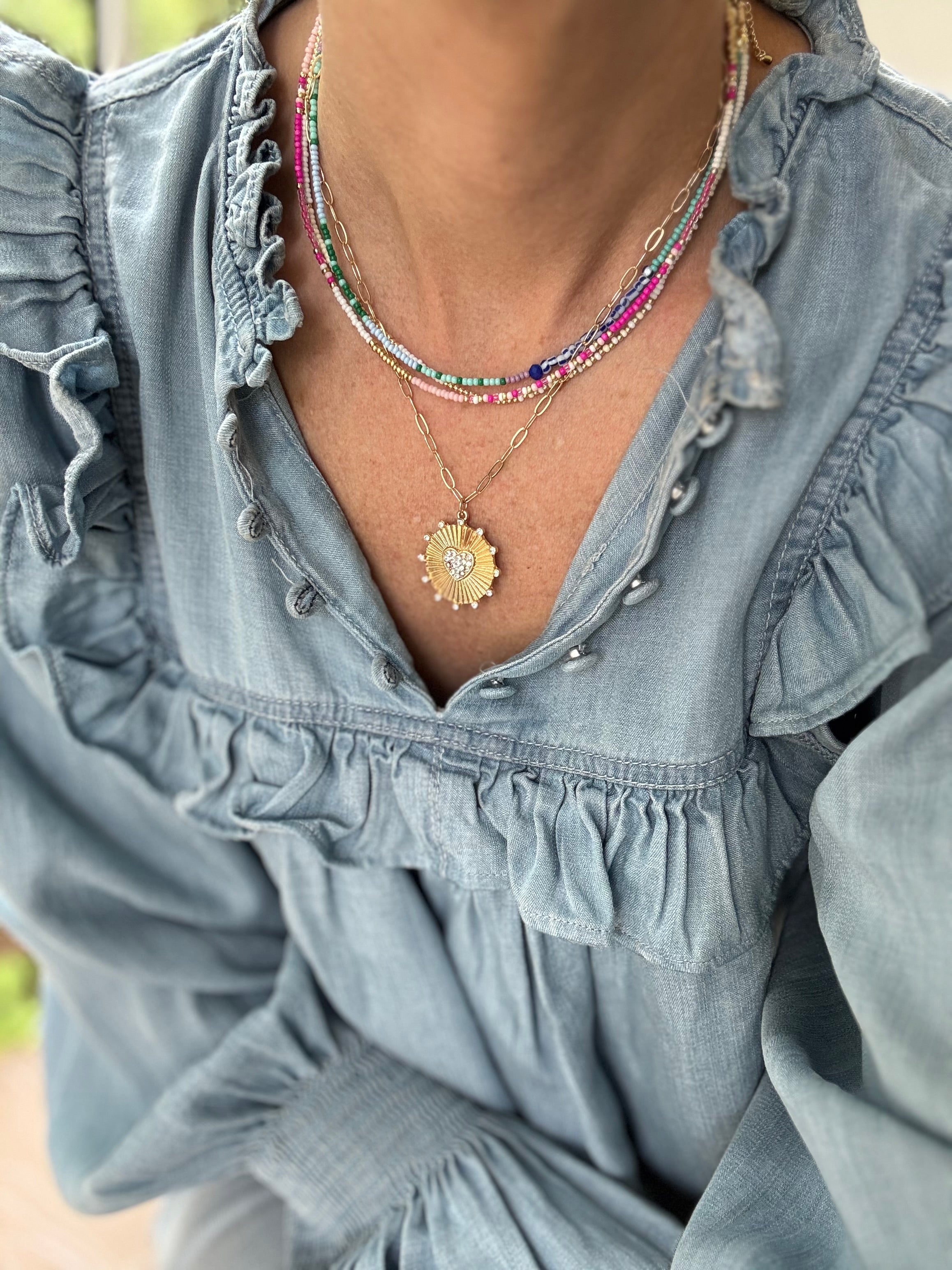 Picture of a woman wearing a blue blouse and four necklaces. 3 necklaces are dainty bead necklaces in pink and white, blue and light blue and white and gold. One necklace is a gold link chain necklace with a large textured gold disc pendant.