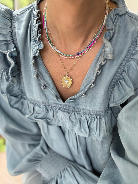 Picture of a woman wearing a blue blouse and four necklaces. 3 necklaces are dainty bead necklaces in pink and white, blue and light blue and white and gold. One necklace is a gold link chain necklace with a large textured gold disc pendant.